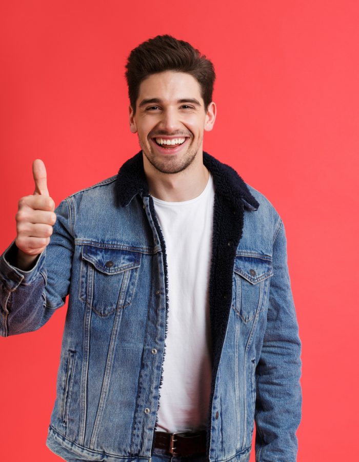 Portrait of a cheerful young man dressed in denim jacket standing isolated over red background, showing thumbs up