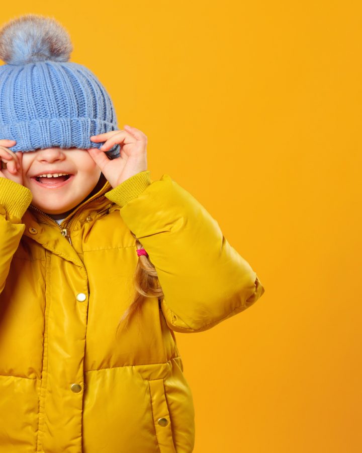 Closeup portrait of a cheerful little girl in jacket over yellow background. The child hides his eyes under the cap.