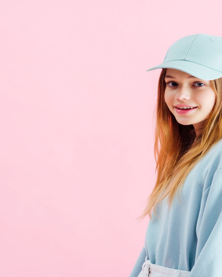 happy pretty teenage girl in cap looking at camera isolated on pink